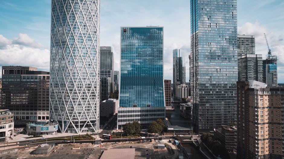 A cityscape view of high-rise buildings in Canary Wharf with modern glass facades reflecting the sky and surrounding structures. In the foreground, there is a section of a well-maintained urban garden