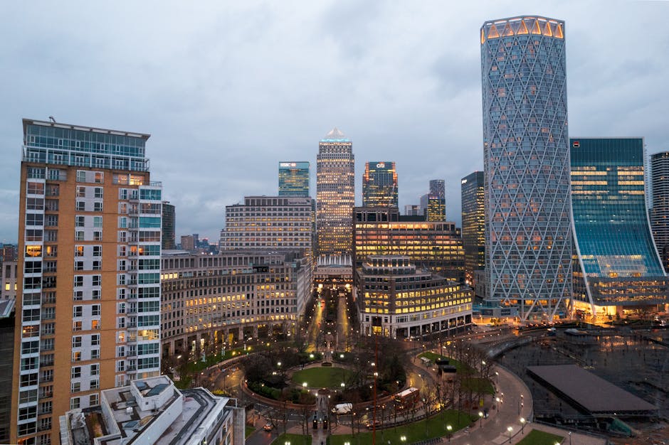 A cityscape view of high-rise buildings in Canary Wharf with modern glass facades reflecting the sky and surrounding structures. In the foreground, there is a section of a well-maintained urban garden