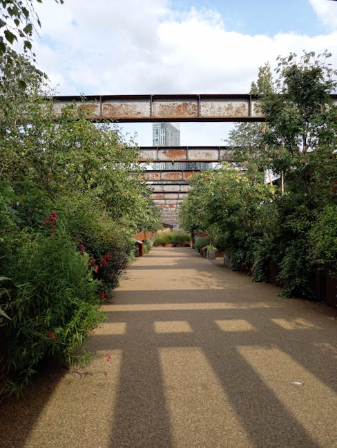 A paved pathway lined with lush, well-maintained shrubbery and flowering plants on both sides, leading towards a modern, industrial-style bridge structure in the background. The garden beds are filled